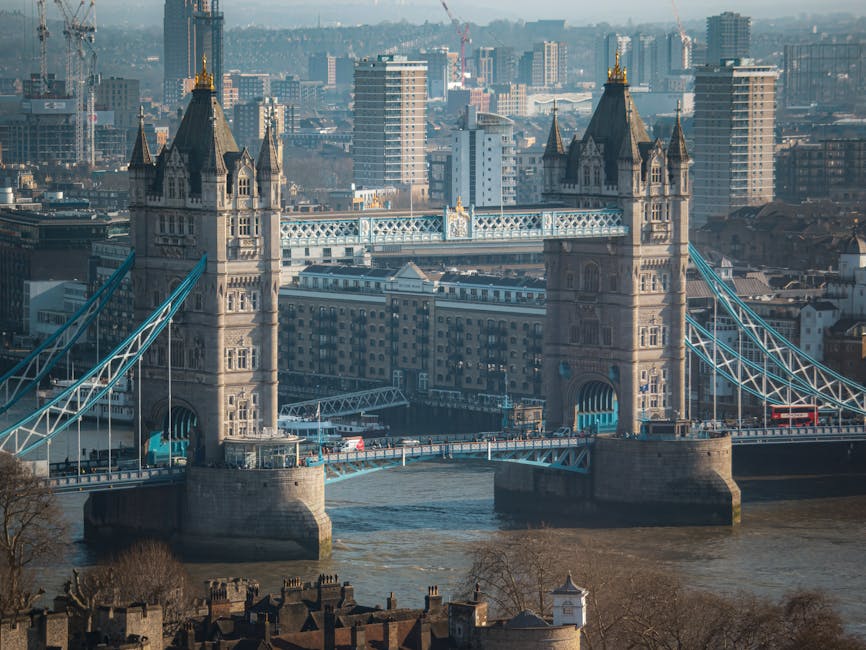 A daytime view of Tower Bridge on the River Thames in London, with its iconic towers and suspension cables prominently displayed. The bridge's structure features stonework and metalwork painted in lig